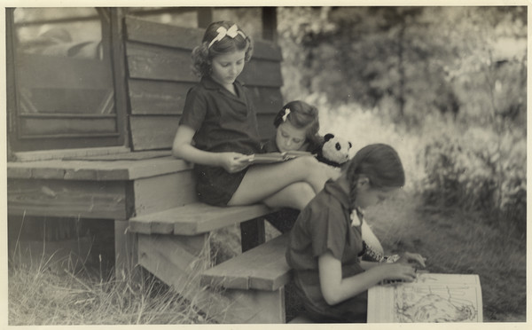 Three campers relax outdoors on the front steps of a cabin. Two of the campers, one holding a stuffed animal, are sharing a book. The third camper, seated on a lower step, is looking at a large book. Original caption notes: "Informal drawing and painting are enjoyed in many leisure moments, especially by the campers in Cabin V. The non-competitive and more leisurely type of program is ideally suited to girls of all ages."