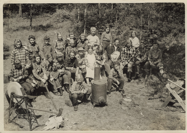 Group portrait of the cast of the Paul Bunyan pageant. All of the campers are in costume, and are arranged in a semi-circle around some of the implements used in the play: a grindstone for sharpening axes, several axes, a woodstove, and some lumber. The group is arranged in a grassy, sunny area with woods in the background.