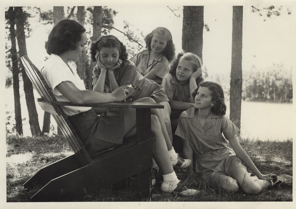 A camp counselor, seated in an Adirondack chair, wearing a white shirt, dark shorts, and saddle shoes, reads to four young campers who are gathered around her. The campers are all wearing the camp uniform. They are posed in a wooded area, and a lake and shoreline are in the background.