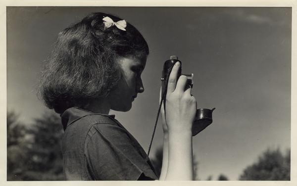Profile view of a camper posing outdoors with a camera, which she is holding up, ready to take a picture. She has a bow in her hair and is wearing the camp uniform. Treetops are in the background. Original caption notes: "Photography also plays a very important part in camp life, for we like to preserve in snapshots the high points and adventures in camp life. This our Camera Club has many active members, both camper and counselor."