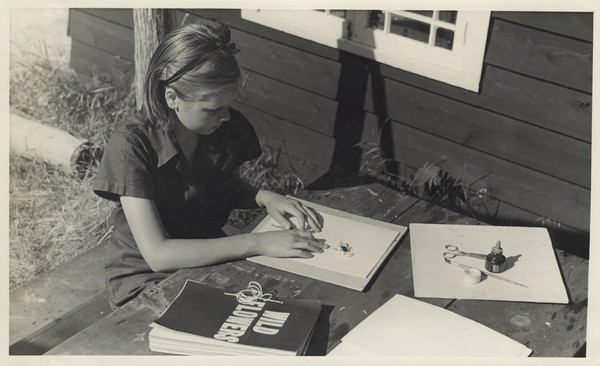 A young camper is seated at an outdoor wood table arranging a flower on a sheet of paper. On the table near her are spread out all the supplies she is using: a hand bound book on wildflowers, paper, scissors, glue, and paint brush. Her hair is pulled back with a hair band and she is wearing the camp uniform. Original caption notes: "Nature study permeates every corner of camp life, for in order to enjoy the outdoors and be comfortable in it, we must know and understand nature around us. Excellent wild flower collections are made by some of the campers."