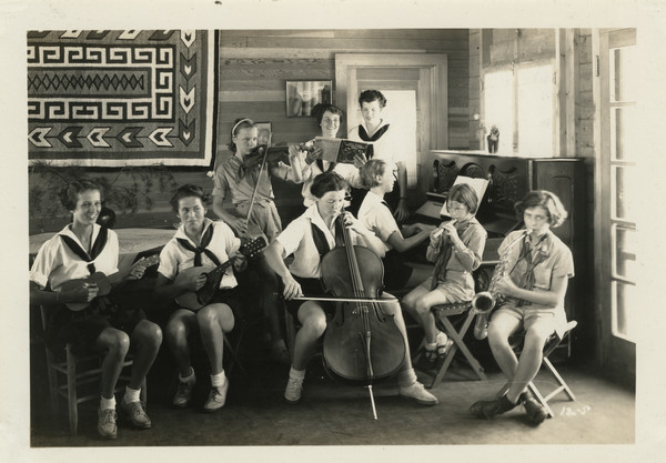 Group of counselors and campers playing musical instruments indoors. Included are several string instruments, a saxophone, recorder, and a piano. Two campers are holding open a songbook. The group is arranged in a corner of an interior room, and a woven wall hanging hangs on a wall behind them. Original caption notes: "Every summer we have at least a dozen campers and counselors whose musical talents add much to the summers' enjoyment for all. As an orchestra, and in trios, duets and solos, those who play instruments find many opportunities to appear during the camp season, especially at our evening musicales and at Sunday Services. Campers who wish to practice on the piano or on a musical instrument may choose to do so, under supervision, if so desired. Music and singing are important and vital parts of our camp life." The caption goes on to note that a graduate student in music was in charge of the music program.