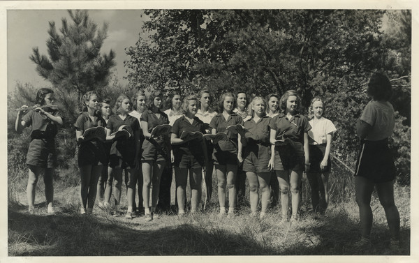 Joy Camps choir, consisting of counselors and campers in camp uniform, posing standing outdoors in two rows. Several campers are holding music books open. A camper to the far left is playing a flute. To the far right a counselor stands with her back to the camera, and appears to be in charge of the group. The choir stands in a sunny, grassy area adjacent to woods. Original caption notes: "Our Camp Choir, under the supervision of a graduate in Music Education, is one of the most enjoyed of all our camp activities. Sunday visitors to our Services will attest to this fact."