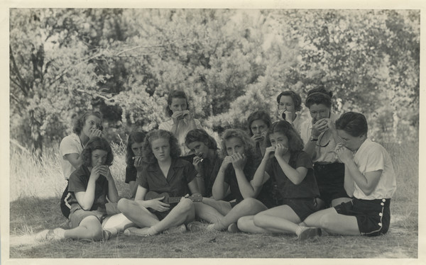 Camp harmonica band posing outdoors in two rows in a shady, grassy area adjacent to woods. Campers and counselors are all seated on the ground with their harmonicas raised to their mouths. One camper plays a small string instrument. Camp owner and co-director, Barbara Ellen Joy, is in the back row, first from the right. Campers and counselors are all wearing their respective camp uniforms. Original caption notes: "In 1939 we organized an Harmonica Band, which was great fun."