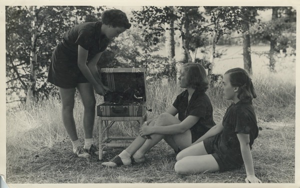 Three campers gathered around a record player on a chair in a grassy area on a hill near trees. One camper is standing over the record player, holding a record. The other two are seated on the ground. All are wearing camp uniforms. Original caption notes: "... always at the disposal of our campers is a splendid collection of fine records. Singing and music in their various forms are enjoyed each day."