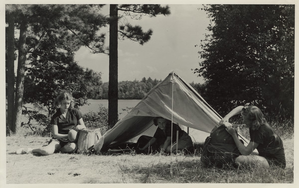Three campers, all wearing the camp uniform, sitting outdoors with small tent. One camper is kneeling in the tent, looking out at the camera. The other two campers are outside of the tent, sitting on the ground. One is sitting with a basket, the other with a backpack. The campsite is located in a grassy area, with the lake and far shoreline in the distance. Original caption notes: "A typical camping out scene. Notice the tents with mosquito tents inside, a promise of perfect sleep. A reflector oven always goes out on our trips. Properly placed opposite a reflector fire, it turns out large batches of golden corn bread, biscuits, shortcakes, and even delicious cake, later to be adorned with chocolate frosting. Campers practice at main camp, as part of the regular daily program, the special skills which are a part of the campers' kit. By canoe, boat, and horseback, in and around our own Lake and all over the surrounding country, we go out on these trips. This knowledge of the outdoor life, and the resulting appreciation of and at-home-ness in nature, are things which campers can always use and treasure, and never forget."