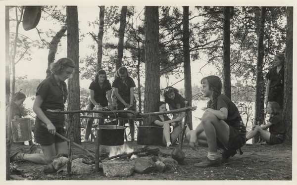 Campers cooking over an open fire. In the foreground, one camper is kneeling on the ground, tending the fire. Two large metal pots hang from thin tree branches arranged over fire. A cast iron skillet hangs above the fire to the left, somewhat out of view. Another camper looks on nearby. A number of other campers are in the background, some standing and some sitting on the ground among trees. The lake and far shoreline are in the distance.
