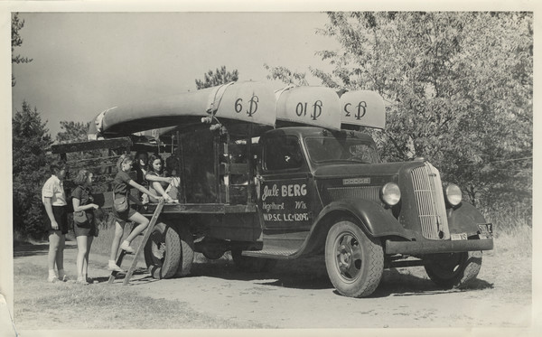 A camper is climbing a short ladder into the back of a Dodge truck where other campers are seated. Another camper waits in line, with a camp counselor looking on. Painted on the passenger side door of the truck are the words: "Jule BERG, Hazelhurst, Wis. W.P.S.C. L.C.#12097." (The last number is partially hidden by a part of the truck.) Three Joy Camps canoes are overturned and fastened to the roof of the truck. Each canoe is numbered and includes the Joy Camps (JC) logo. The truck is parked on a sandy road near trees.