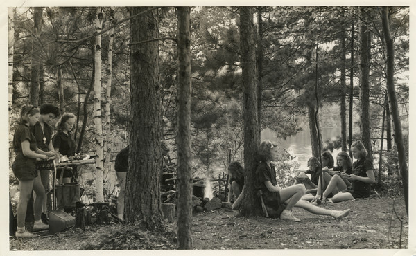 Group of campers under trees near a lake eating from metal dishes next to a campfire. A few of the campers are relaxing, and some appear to be washing the metal dishes used in the meal. The campfire is in the center, somewhat hidden by tree trunks. Camping utensils are scattered about the site, including some tin cups hanging from a tree trunk and an ax resting against another tree trunk. Two other campers are out on the lake in a canoe, approaching the shoreline.
