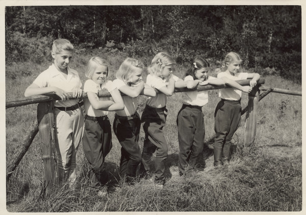 Several younger campers leaning against wood fence, near wooded area. They all appear to be wearing jodhpurs.