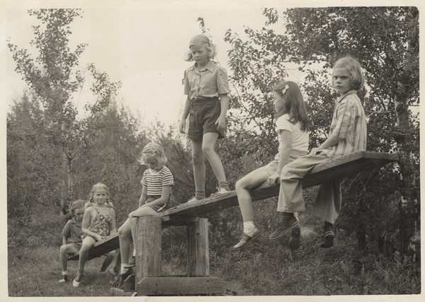 Group of younger campers sitting and standing on a wood seesaw near trees.