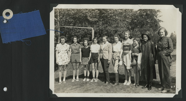 Page from Joy Camps photo album displaying a group portrait and a swatch of blue cloth from the official camp uniform. Original caption notes: "Our camp outfits are as simple and inexpensive as they possibly can be. Parents are urged to send items from the regular home supply in so far as possible. From left to right: any type of play, halter or gym suit; 'official blue' uniform and light-weight sweater (Carson's outfit us with these practical and inexpensive blue suits); Sunday uniform of white blouse or shirt and dark blue shorts; riding outfits of any sort; swim suits of any type; rain togs consisting of rubbers or rubber boots (which are very practical), sou'wester or rain hat, and slicker; ordinary boys' overalls or jeans, leather belt, and — VERY important to all Joy Campers — a red or blue bandana."