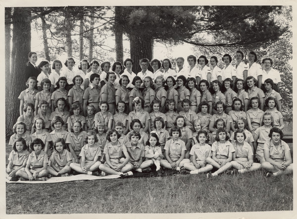 Outdoor group portrait of campers and counselors during one season (undated). Original caption notes: "The entire group during one of the periods the past season. Counselors are wearing white shirts, the campers the regulation blue costume. Seldom will you see a healthier, finer, happier, or more wholesome group of people." Back of photo carries stamp: "Minocqua Photo Shop, Victor A. Hendrickson, Minocqua, Wis."