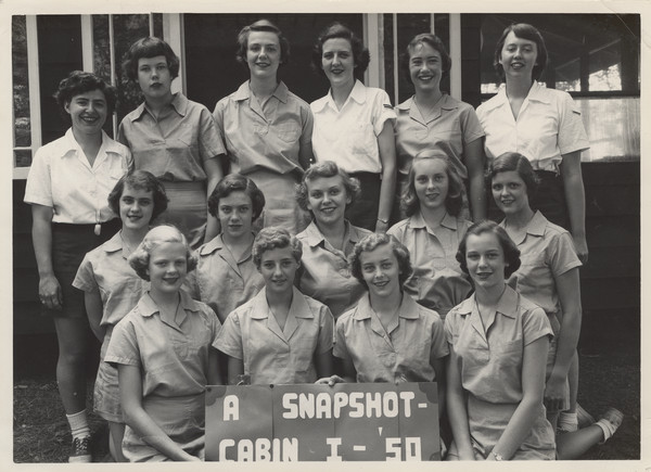 Outdoor group portrait of older campers, with counselors, posed in front of a camp building. The girls in the front row are holding a sign that reads: "A Snapshot, Cabin I — '50." Sue Ann Hackett, the person who donated these photographs, is in the front row, second from the right. Original caption notes: "The oldest of the three Cabin groups which make up Deerpath Lodge for older girls, those of junior and senior high school age. Our flexible, non-schooly program appeals to girls of ALL ages, but especially to those who are older and able to think for themselves. The days are too short for them to do all the things their advanced skills and interests make possible for them to achieve. Many of these girls will soon be going to colleges, where they will find their camp experience and associations of great value and enjoyment. Loyal and enthusiastic campers, these girls return home after camp healthy and fit, ready for the strenuous months ahead." Back of photograph carries stamp: "The Joy Camps, Hazelhurst — Wisconsin" and "Minocqua Photo Shop, Victor A. Hendrickson, Minocqua, Wis."