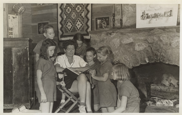 Interior view of group near the Main Lodge fireplace, with Barbara Ellen Joy seated in a camp chair reading to a group of young campers. Numerous decorative items are shown: a record player, a small drum, a print on either side of a woven rug, all hanging on the wall, and another print and two candlesticks on the mantle of the fireplace. Original caption notes: "Miss Joy and members of Camp Pioneer. Both directors keep in close and friendly touch with each camper and the day-light hours of both are spent actively with the campers and their leaders."