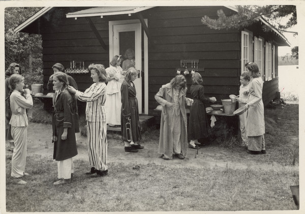 Large group of campers outside their cabin, dressed in pajamas and bathrobes. Some of them are using dippers with metal pails; on the far left a row of dippers are hanging from brackets on the exterior siding of the cabin, and toothbrushes and cups hang from brackets on the right. One girl is braiding another girl's hair, another girl is carrying a bugle, and two girls standing near the screen door to the cabin (one inside, one outside) are holding a doll and a stuffed animal (toy) rabbit. A lake and distant shoreline are in the background on the right. Original caption notes: "Cabin IV getting ready for bed!"