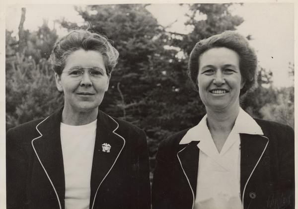 Outdoor quarter-length portrait of Joy Camps co-directors, Barbara Ellen Joy (on the left) and Marjorie Camp (on the right). Both women are wearing white shirts and matching dark jackets. Miss Joy also has a pin attached to one of the lapels of her jacket. A wooded area is in the background. Back of photograph carries stamp: "Minocqua Photo Shop, Victor A. Hendrickson, Minocqua, Wis."