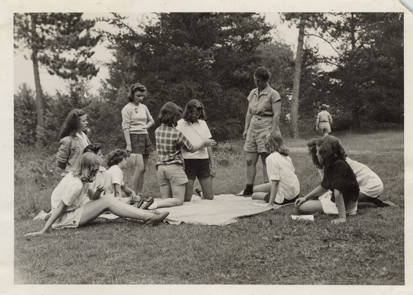 Two campers kneel on a blanket on the ground, demonstrating bandaging. Several other campers are gathered around the blanket, some on the ground, some standing. Camp co-director, Miss Camp, is also in the group. Original caption notes: "... Miss Camp and her class in another phase of 1st Aid, bandaging."