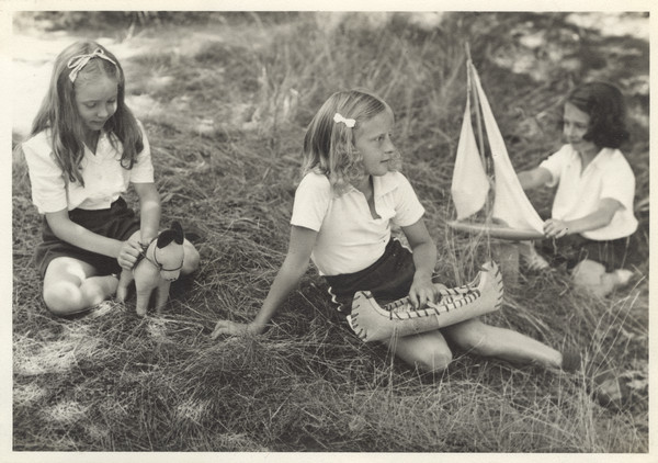 Three young campers sit in a shady, grassy area, each with a toy. One has a stuffed animal horse, one has a toy canoe, and one has a toy sail boat. All three campers are wearing white shirts and dark-colored shorts. Original caption notes: "Three Cabin 'V'ers of 1943 with their toys."