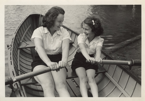 Close-up of a camp counselor and young camper in a rowboat on the water. They are looking at each other and smiling. Both are wearing white shirts and dark-colored shorts.