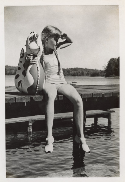 Close-up of a young camper sitting on a dock wearing a swimming suit, and holding a floating device shaped like a fish or seahorse. She has her hair in two braids and is wearing open-toed shoes with straps. The far shoreline is in the background.