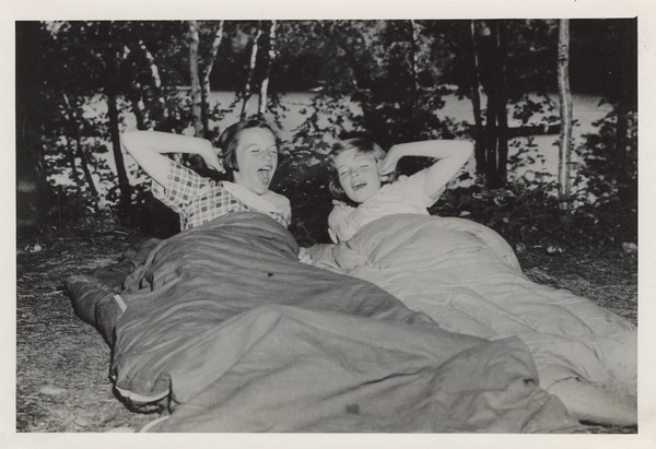 Two campers yawn and stretch from their sleeping bags near a shoreline. Trees, the lake, and far shoreline are in the background.