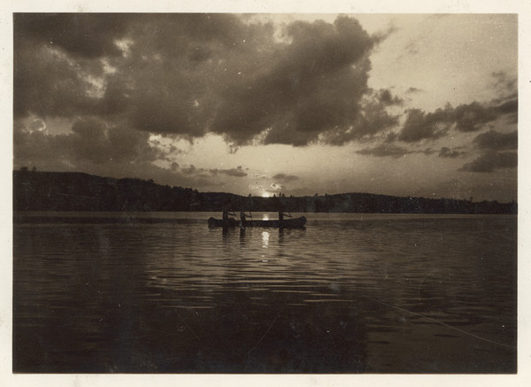 View across water towards three campers in a canoe who are silhouetted against a sunset over a lake. The tree-lined shoreline is in the far background.