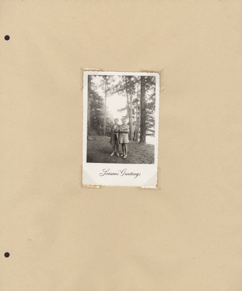 Page from Sue Ann Hackett Blue Album displaying a holiday photograph, with co-directors Miss Camp on the left and Miss Joy on the right. They are dressed casually, standing near trees. Written below: "Season's Greetings."
