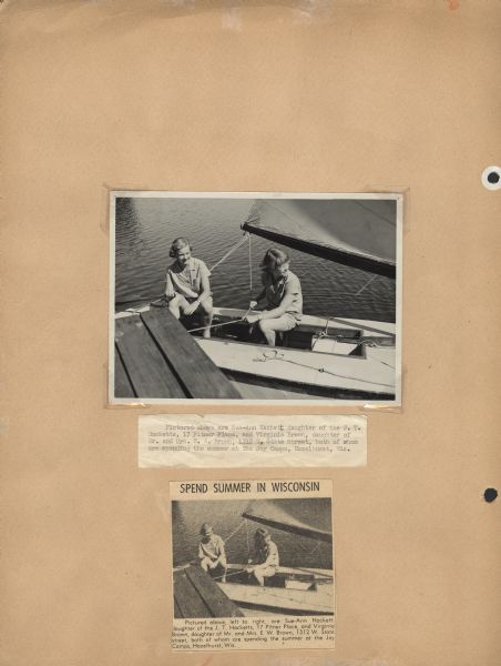 Page from Sue Ann Hackett Scrapbook displaying a newspaper clipping with photograph and caption; Sue Ann Hackett and friend Virginia Brown are pictured here on a sailboat alongside a dock. The caption notes that "both ... are spending the summer at the Joy Camps, Hazelhurst, Wis." The original photograph used in the newspaper clipping is included on this album page along with a typewritten caption. Both girls are wearing the camp uniform and are handling different parts of the sailboat; Virginia Brown is wearing glasses.