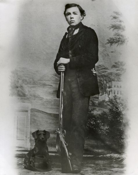 Studio portrait in front of a painted backdrop of a young Paul A. Seifert. He is posing standing and holding a rifle, and a small dog is sitting at his feet behind a hat resting on the floor.