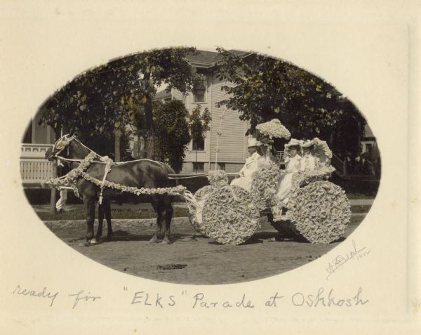 Hand-colored view of a horse and carriage decorated for a parade. Jessie Jack Hooper, wearing a floral hat, holds a decorated parasol in the front seat. A man, probably Benjamin Hooper, wears a white suit and hat and holds the reins of the horse. Lucille Peck and Lillian Hooper, both in floral hats, hold decorated parasols and ride in the back seat.
