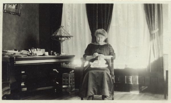Jessie Jack Hooper sitting in a rocking chair at her home at 669 Algoma Street in front of two windows with heavy drapes and lace curtains. She is stitching a garment she holds in her lap. A table to Ms. Hooper's right is covered with books, papers, and inkwells. There is also a lamp on the table.