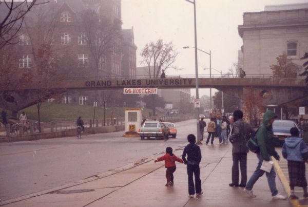 Park Street at the State Street intersection during the filming of <i>Back To School</i>. The pedestrian overpass bears a sign for the fictional Grand Lakes University and a banner reading "Go Hooters." There is an information booth set up in the middle of the street where two cars are stopped. Several pedestrians mill about on the sidewalk. The Wisconsin Historical Society headquarters building is on the right. Science Hall is in the upper left and Helen C. White Hall is at center behind the overpass.