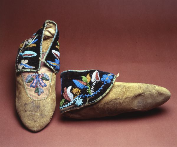 Men's buckskin moccasins. Puckered toes with purple cloth flaps that are patially decorated with floral pattern in seed beads. Sewn with the spot stich, black velvet cloth flaps decorated similarly and edged with green ribbon. Blue, green, pearl colored beads. Tongue is purple. Center seam meets toe point.