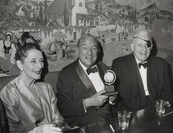 Lynn Fontanne, Sir Noel Coward, and Alfred Lunt seated at a table in the Pyrenees Restaurant after the Tony Awards presentations at which Lunt and Fontanne received the Antoinette Perry Award for Excellence. Mr. Lunt wears an eye patch due to the loss of sight in his left eye. The group is seated in front of a mural of a French scene.