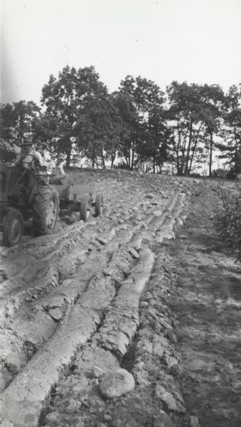 Alfred Lunt driving a tractor pulling Ben Perkins in a cart. Mr. Perkins is likely seeding the furrows.