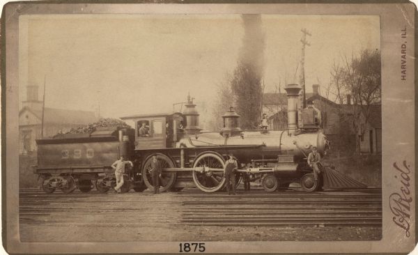 View across railroad tracks towards a small group of men posing with Chicago & Northwestern Railway locomotive number 390 and a coal car. Dwellings and a church are in the background.