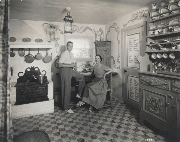 Alfred Lunt stands at the edge of a table holding a beverage, near Lynn Fontanne who is sitting, in the kitchen of the cottage at their Ten Chimneys estate.