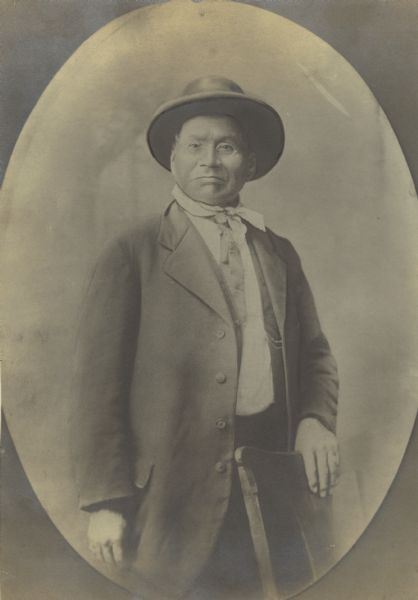 Three-quarter length formal portrait in front of a painted backdrop of John Nuwi, a Mascouten (or Prairie) Potawatomi, who lived at Skunk Hill in Wood County, Wisconsin. Powers Bluff County Park, site of Skunk Hill, is on Bluff Drive, one mile west of County E in the town of Arpin, about midway between Wisconsin Rapids and Marshfield.

