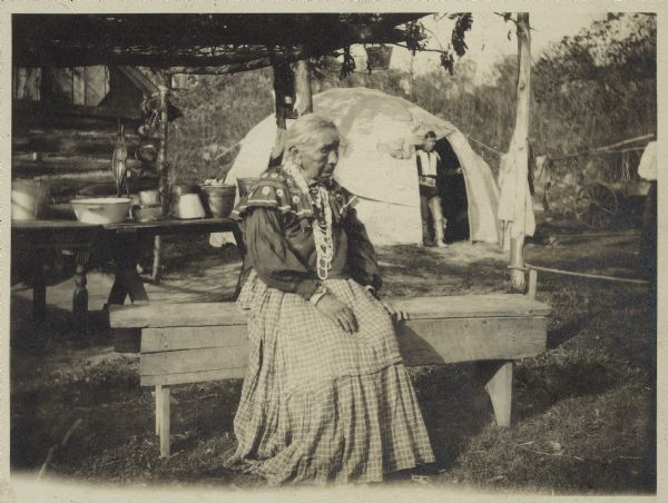 Informal portrait of a Potawatomi woman, Kikyako from Phlox, sitting on a bench wearing a beaded necklace. A man is standing in the doorway of a wigwam in the background.