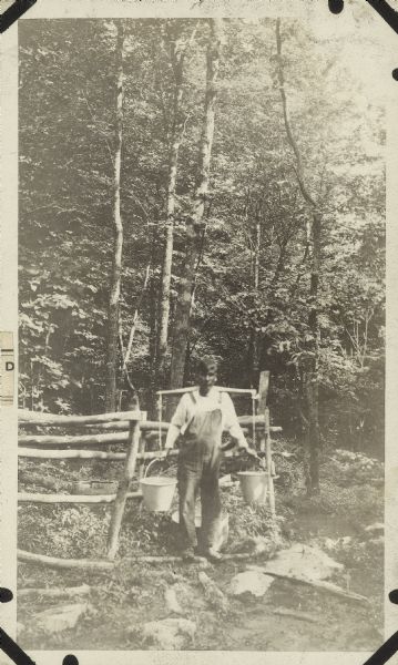 Outdoor portrait of a man standing near a spring in a wooded area surrounded by a wooden fence. He wears coveralls and holds two pails with a yoke over his shoulders for carrying water.