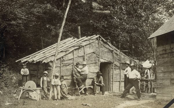 Small group of men and boys gathered outside a traditional elm-bark structure at the upper part of the Skunk Hill community. A young boy stands on the support frame of a whet-stone on the left. The side of a wood building with a roof is on the right.