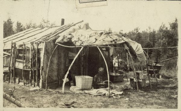 Anna Goodvillage, at her home at Hemlock Creek, weaving a basket while sitting beneath a wigwam type shelter attached to the side of a wood and bark frame structure.