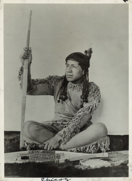 Portrait of Chicog seated cross-legged on the ground holding a bow. There are snowshoes in the foreground.