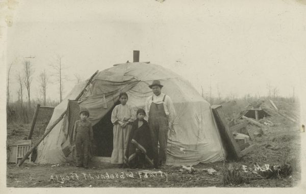 Group portrait of Albert Thunder, his wife and two children standing outside a canvas covered wigwam.