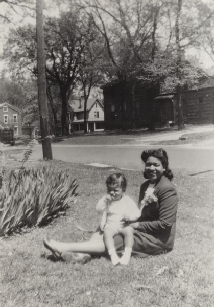 Frances Reneau with her son Thomas on her lap seated on a lawn on a residential street. Mrs. Reneau wears a corsage on her lapel and the toddler, Thomas, holds a flower.