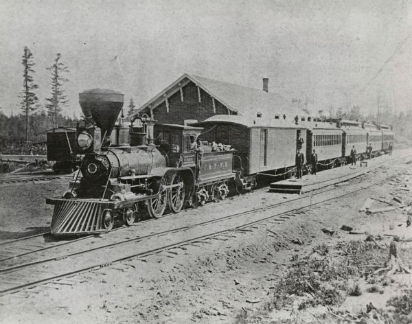 Slightly elevated view of a Chicago & Northwestern Railway train at a depot in an unidentified town. The railroad station is behind the train. On the right, porters stand on the platform at the entrances to the passenger cars. The engineer and another man lean from the engine. Note the deer head mounted to the front of the stack.