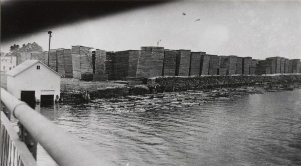 View from bridge or boat railing of piles of lumber in the Paine Lumber Company yards on the banks of the Fox River. There is a boat house on the left shoreline, and a commercial building is behind it. A log raft is along the shoreline near the stacked lumber.