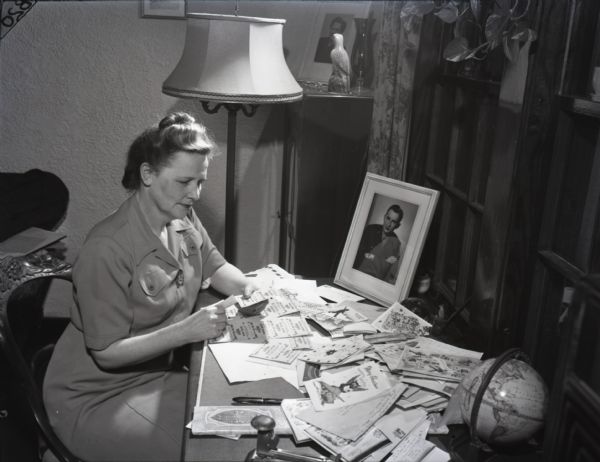 Informal portrait of Rosina Victoria Junghans seated at a desk with cards and letters to and from servicemen and women serving in World War II. She holds ration coupons which, presumably, she sent as gifts to pen pals serving in the military. There is also a portrait of her son, Richard Junghans in uniform, and a rotating globe on the desk.