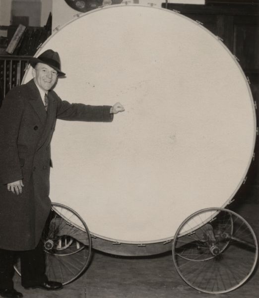 Man in a long coat and hat standing with the large Paul Bunyan drum, which is mounted on wheels.